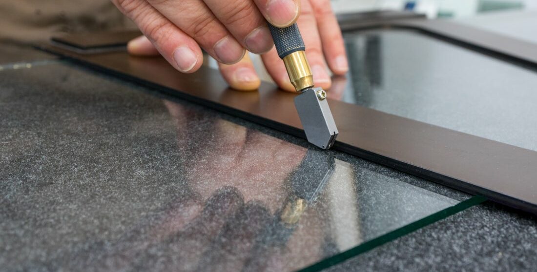 A hand uses a tool to cut down a sheet of glass for repair.