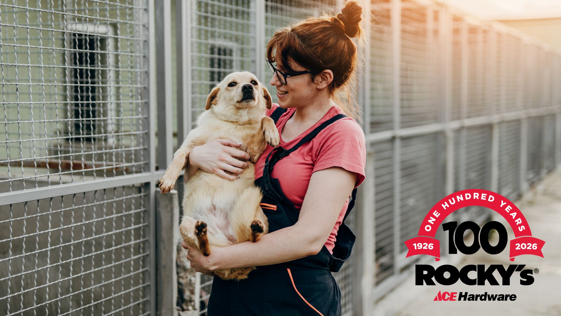 RockysPRJun2026 A pet shelter employee holding a rescue dog at a pet shelter. The Rocky's Ace Hardware 100-Year Anniversary logo is in the lower right corner.