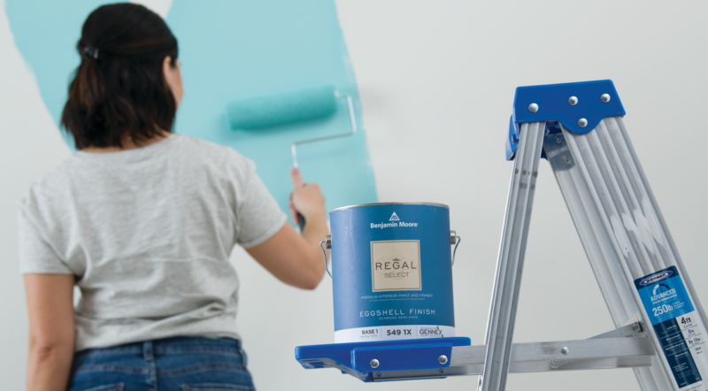 Woman looks at blue paint sample on wall. a Benjamin Moore can sits on a ladder.