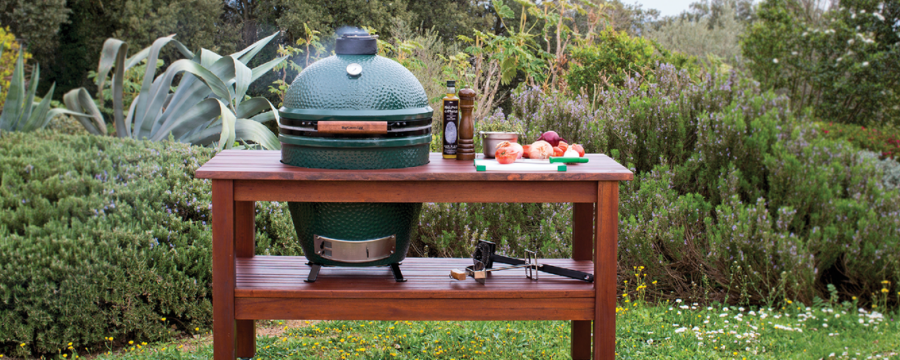 Large Big Green Egg on table with smoke coming out from the sizes. Greenery in the background wit food grilling.